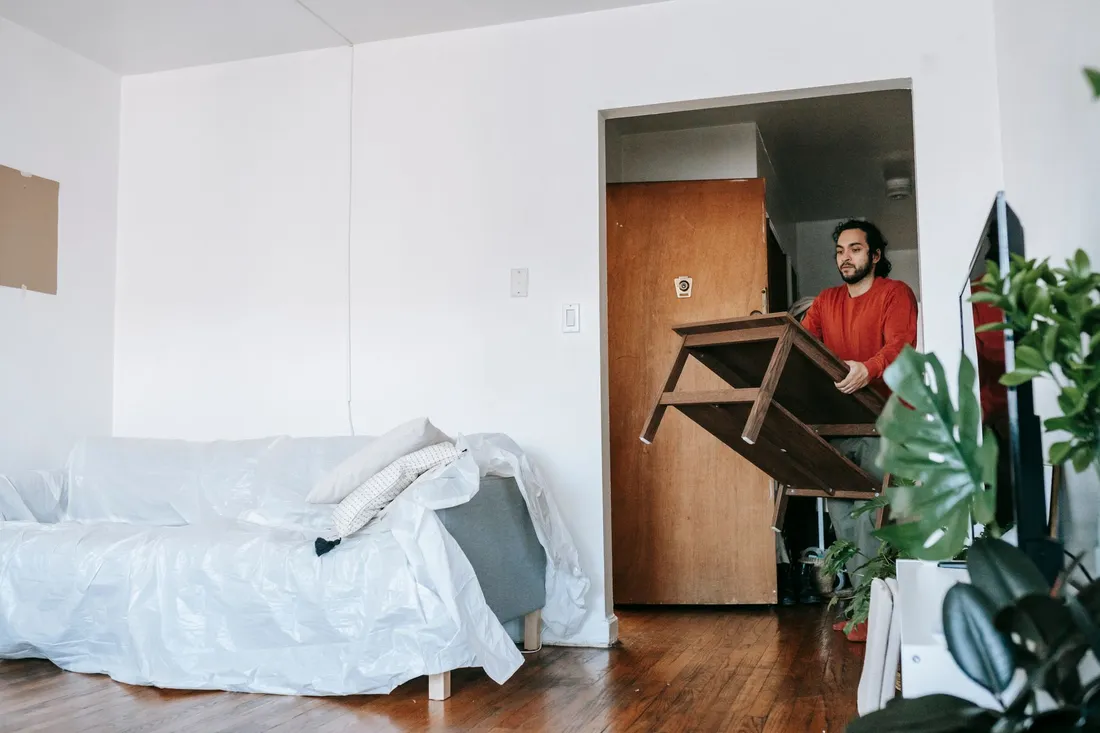 Mann trägt einen Holztisch durch die Wohnungstür, das Sofa daneben ist abgedeckt