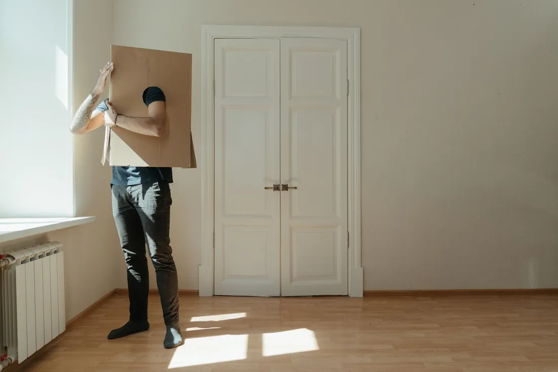 Mann trägt einen großen flachen Karton durch ein leergeräumtes Zimmer mit Parkett